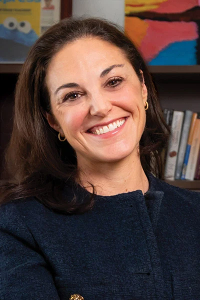 Broadly smiling woman with dark hair in a navy jacket with gold buttons, arms crossed, in front of bookshelves.